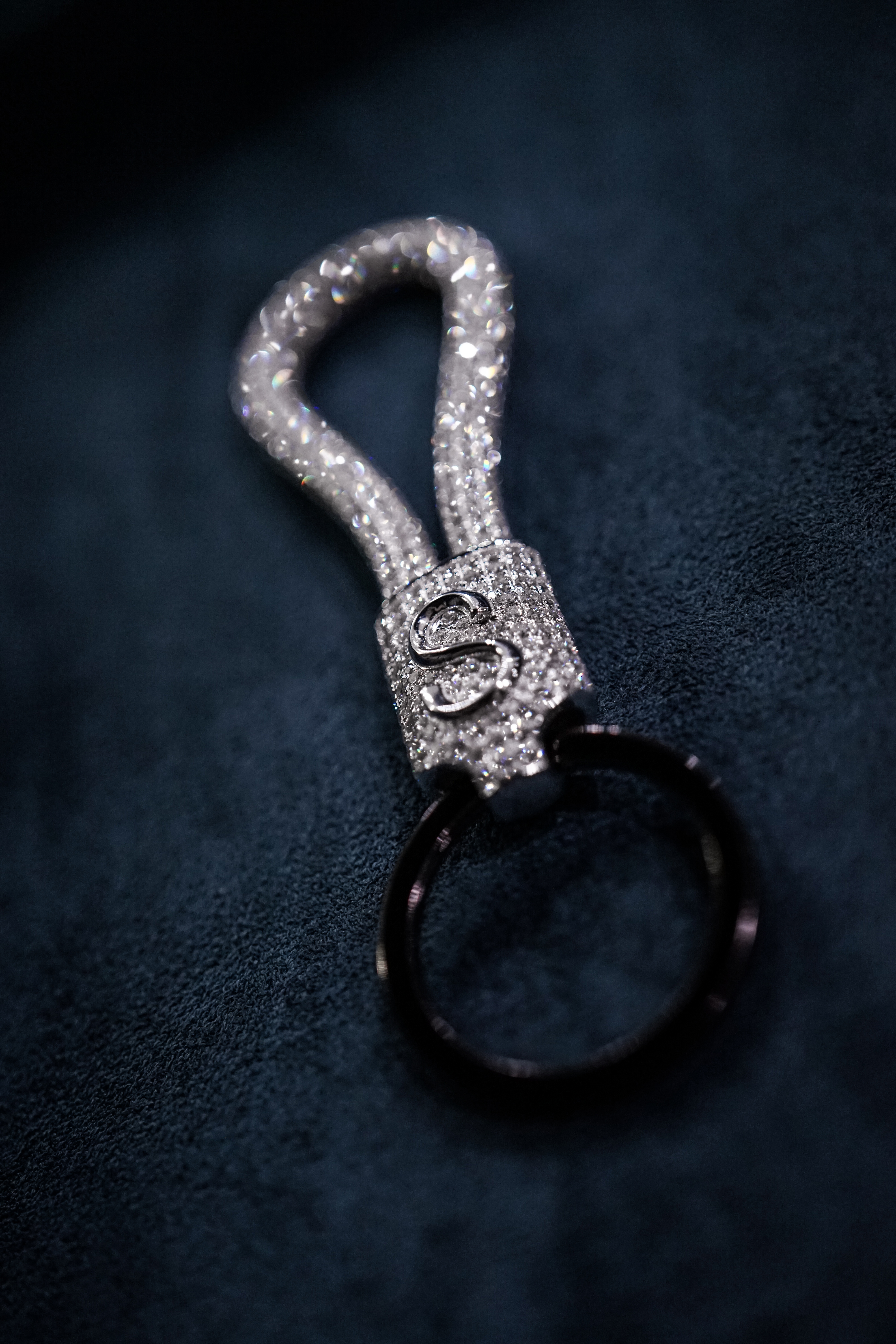 Close-up of a decorative keychain featuring a sparkling, diamond-encrusted loop with a metal clasp. The background is a soft, dark fabric.