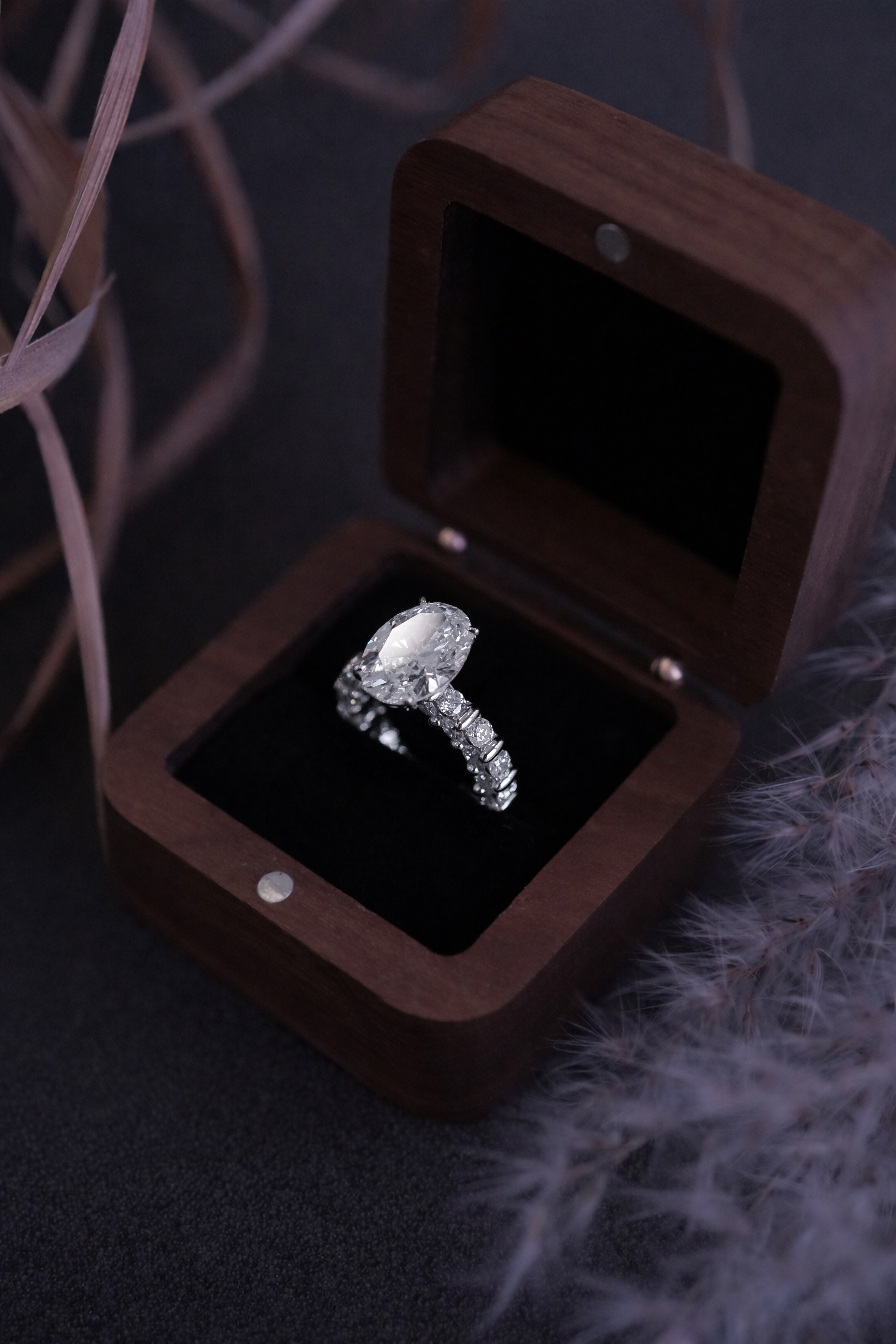 A silver engagement ring with a marquise cut diamond is displayed in an elegant wooden box with a black interior, surrounded by soft, light-colored foliage.